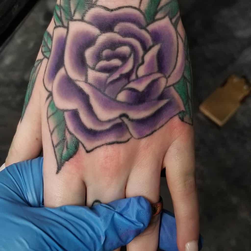 A colorful purple rose tattoo on the hand, with green leaves and soft shading, being worked on by an artist