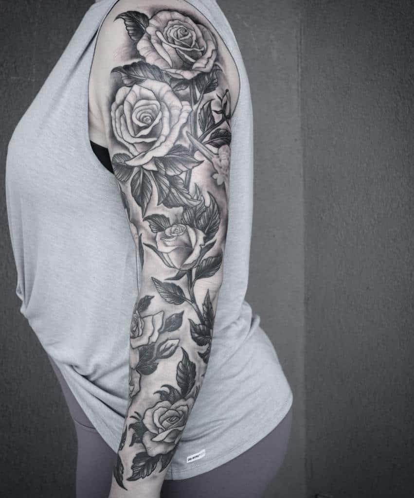 A stunning sleeve of detailed black and grey roses entwined with delicate leaves beautifully covers the woman's left arm