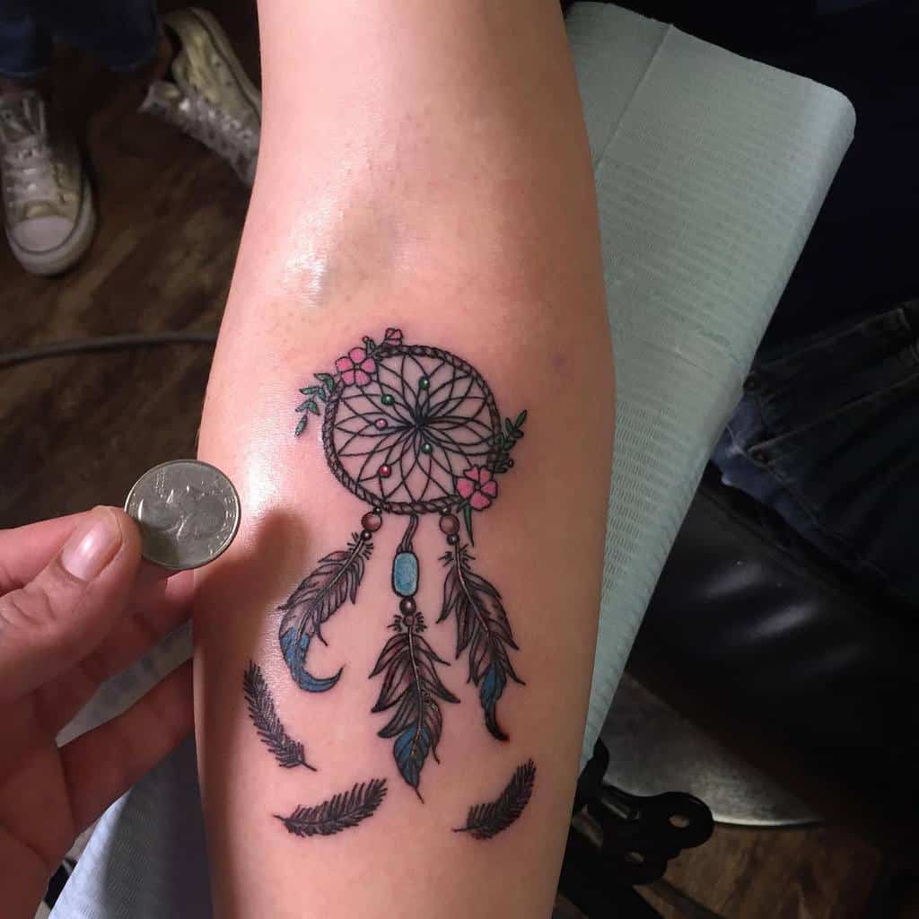 Delicate dreamcatcher tattoo with intricate webbing, pink floral accents, blue beads, and detailed feathers