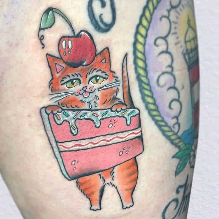 Playful tattoo of an orange cat drooling inside a pink frosted cake with a cherry on top, done in a colorful, cartoonish style