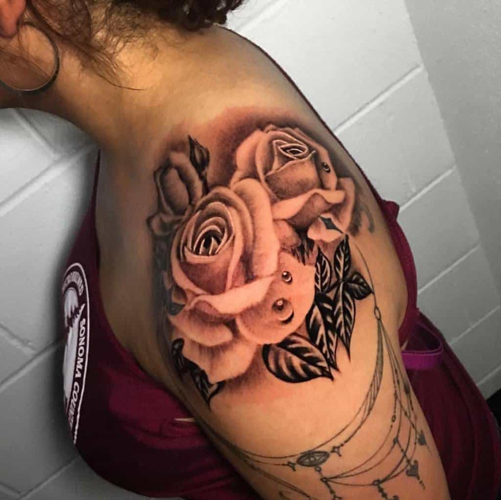 A shoulder tattoo adorns a woman's arm with two large roses in black and gray, complemented by intricately detailed leaves
