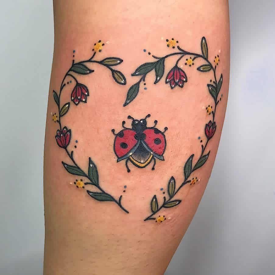 Colorful ladybug tattoo with open wings, framed by a delicate floral heart design with red flowers and green leaves for a whimsical touch.