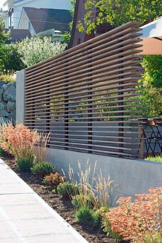 Modern slatted fence with decorative grasses and vibrant plants along a concrete pathway.