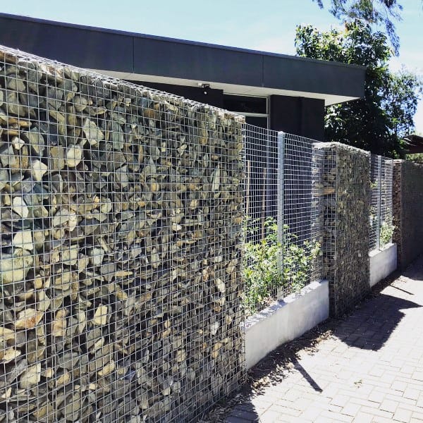 Gabion fence filled with small stones, lining a modern property with a paved walkway.