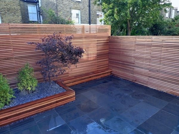 Narrow horizontal wooden fence enclosing a modern patio with raised planters and stone flooring.