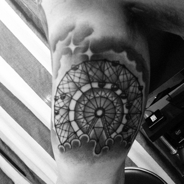 Black and grey Ferris wheel tattoo with bold shading, surrounded by clouds for a dreamy, atmospheric effect on the upper arm.