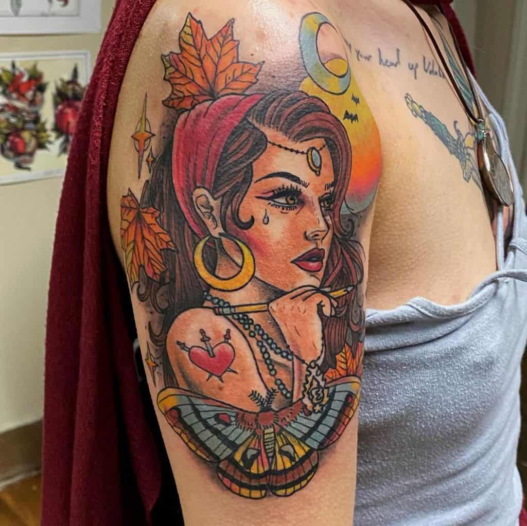 Colorful tattoo of a gypsy woman with a red headscarf, gold jewelry, autumn leaves, a heart tattoo, and a detailed moth on the upper arm.