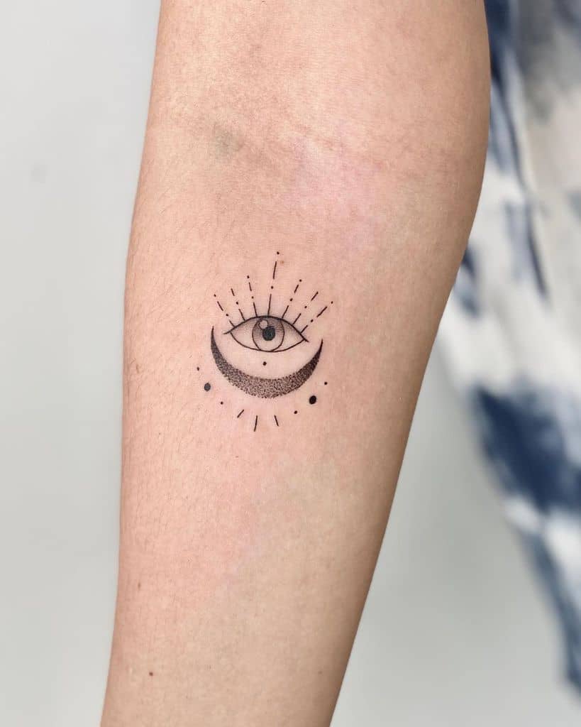 Minimalist tattoo of an all-seeing eye above a crescent moon, surrounded by radiating lines and dots, inked in fine black lines on the forearm.