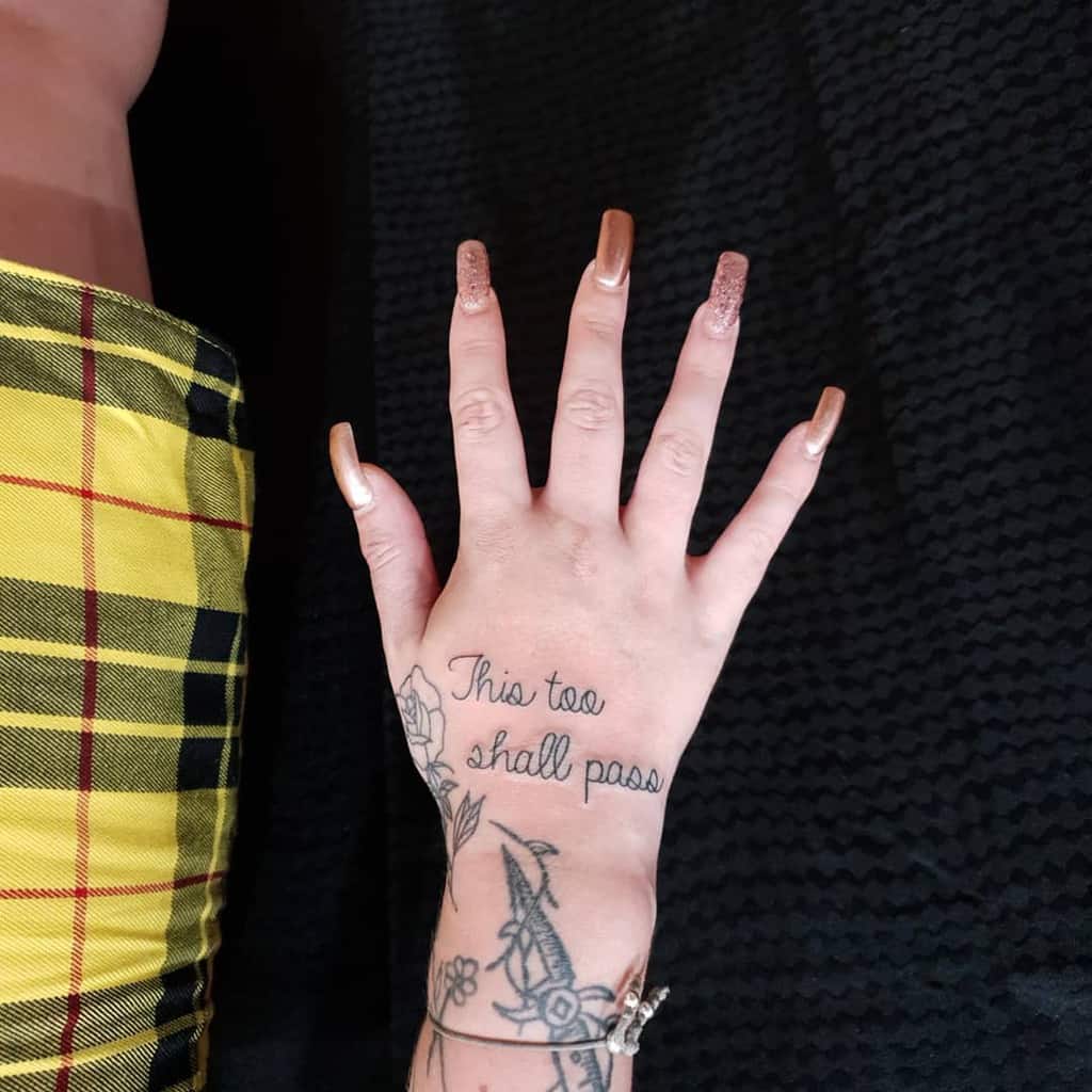 The tattoo on the hand reads "This too shall pass" in a delicate script, accompanied by a small floral design beneath the phrase.