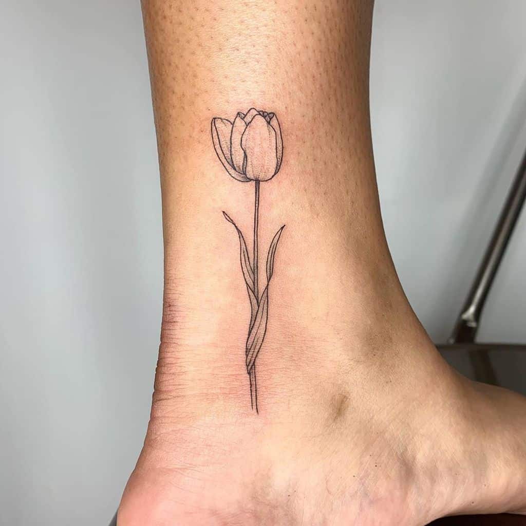 A delicate black line tulip tattoo on the ankle, featuring a slender stem and soft, elegant petals.
