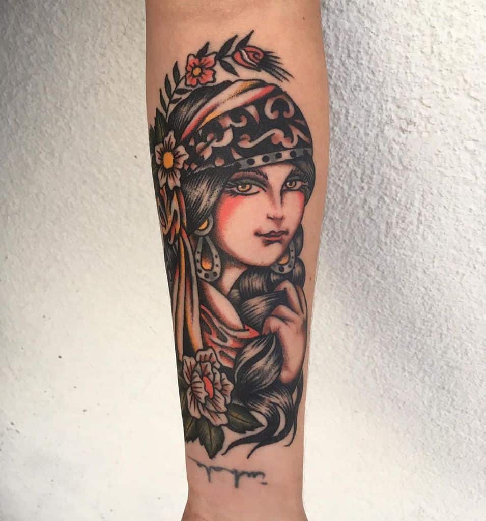 Traditional tattoo of a gypsy woman with a patterned headscarf, gold jewelry, and floral accents, featuring bold shading on the forearm.
