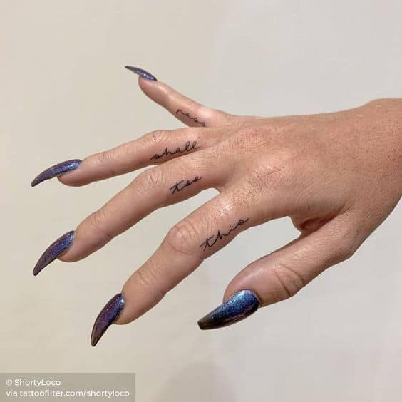 This image showcases a hand with multiple tattoos on the fingers. The tattoos read "this too shall pass" across the fingers in a simple, clean font, and the hand is adorned with long, dark, glittering nails.