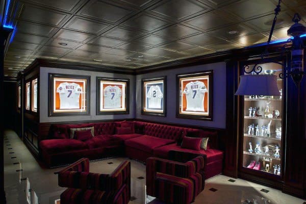 Sports-themed basement with framed jerseys, tufted red sectional, and decorative coffered ceiling.