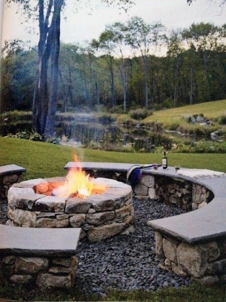 Stone fire pit with wooden benches by a pond, surrounded by trees and grass under an evening sky