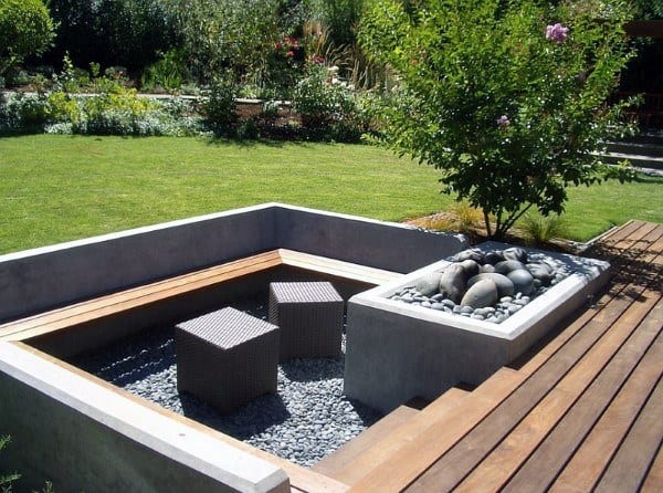 Modern garden seating area with wooden benches, stone pebbles, a fire pit, and two cube stools next to a green lawn