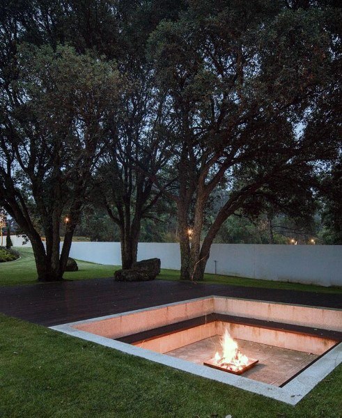 A modern sunken fire pit nestled among trees on a stone patio