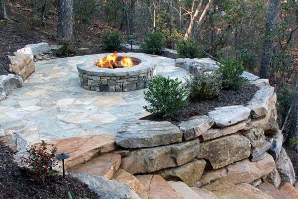 Stone patio with a central fire pit, surrounded by trees and landscaped with bushes and rocks
