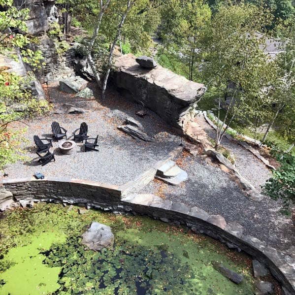 Aerial view of a rocky patio with chairs around a fire pit, near a garden pond with green algae