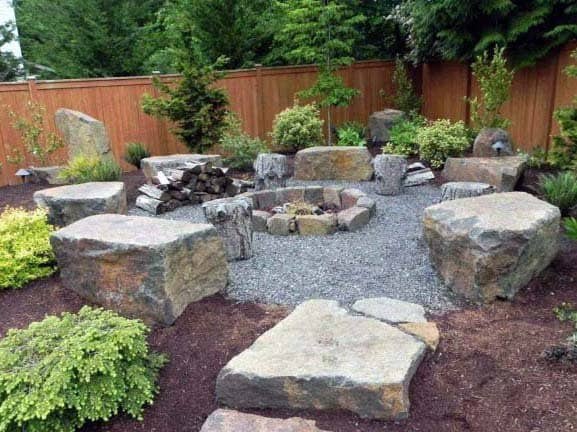 Stone fire pit surrounded by large rocks in a gravel area, enclosed by a wooden fence with greenery
