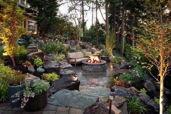 A cozy outdoor patio with a fire pit surrounded by trees, plants, and seating under a serene evening sky