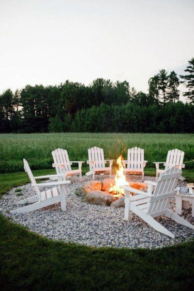 White Adirondack chairs surround a fire pit on gravel, nestled in a lush green field with trees in the background