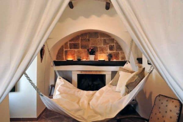 Cozy hammock-style hanging bed with cushions, set near a rustic fireplace under soft lighting.