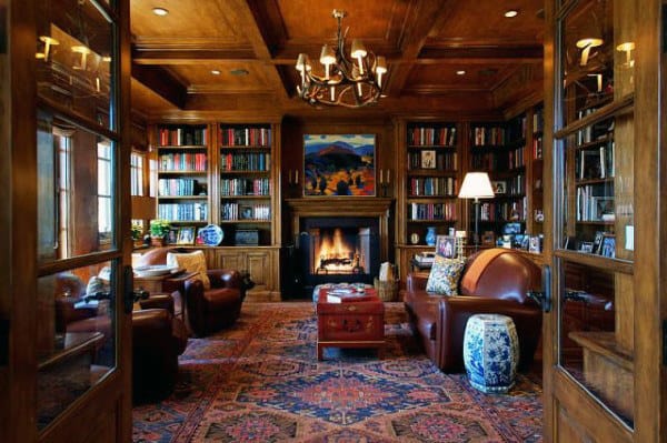 Charming library with wooden shelves, leather chairs, a fireplace, and a colorful rug under a gleaming chandelier