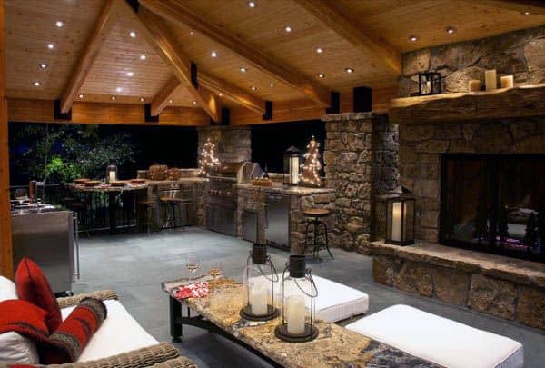 Cozy outdoor kitchen with stone fireplace, wooden ceiling, and seating area warmly lit by lanterns and outdoor string lights
