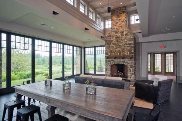 Modern open-concept room with tall stone fireplace, large windows, and rustic wooden dining table.