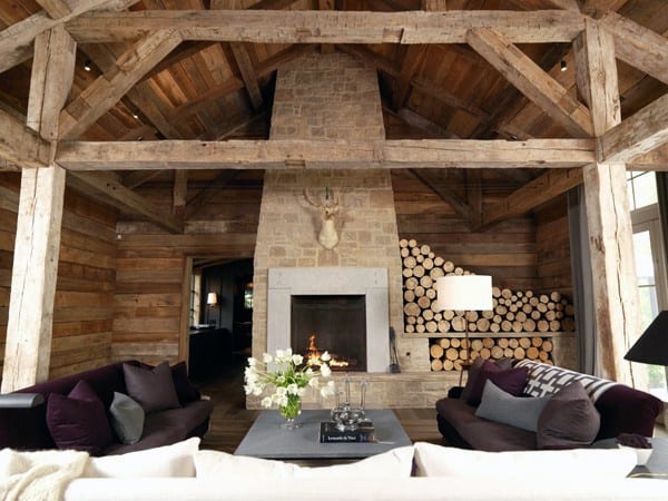 Rustic living room with stone fireplace, exposed wooden beams, and stacked firewood storage.