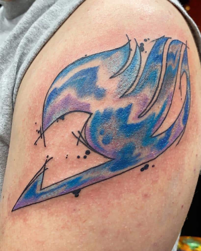 Watercolor-style Fairy Tail guild emblem tattoo on the upper arm, featuring vibrant blue and purple hues with artistic ink splatter details.
