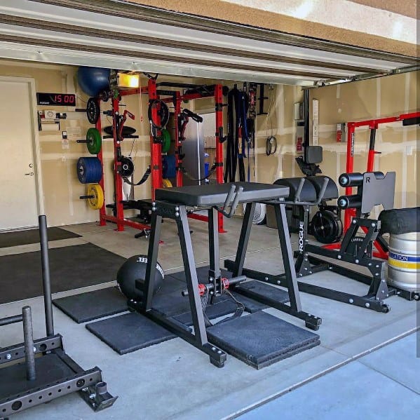 Garage gym with red power rack, GHD machine, sled, weight plates, and organized fitness accessories.