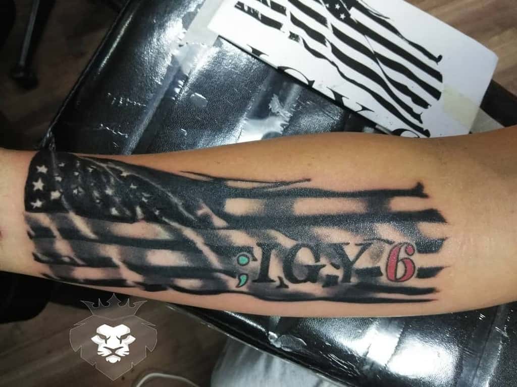 Tattoo of an American flag with "1776" and a semicolon, on an arm in black and gray with some red shading