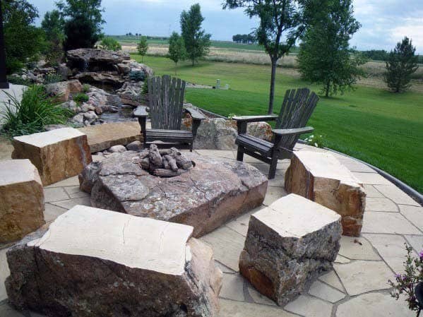 Fire pit surrounded by stone benches and wooden chairs on a patio, overlooking a grassy landscape with trees