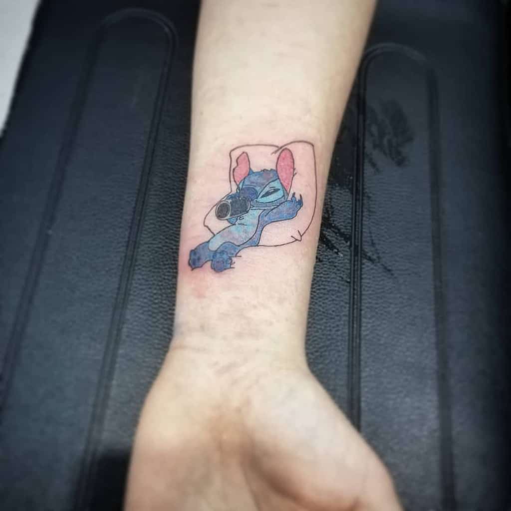 Minimalistic tattoo of Stitch from Lilo & Stitch in a relaxed pose, with soft lines and light coloring, on the forearm.