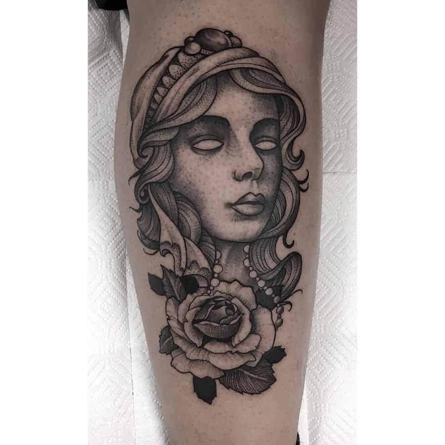 Black and grey tattoo of a mystical gypsy woman with blank eyes, a beaded headscarf, and a detailed rose on the leg.