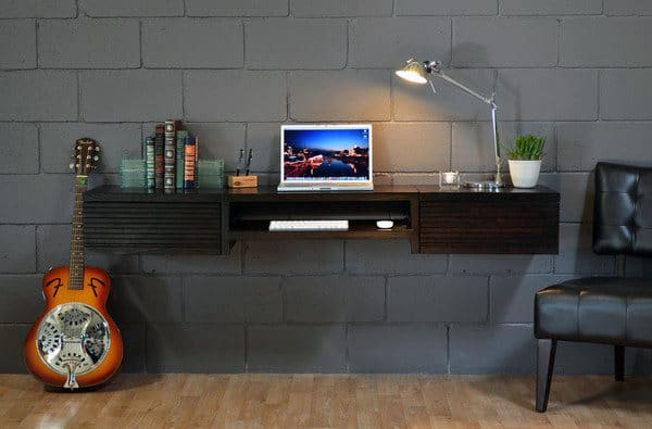Floating desk with a laptop, lamp, books, plant, and guitar against a gray brick wall