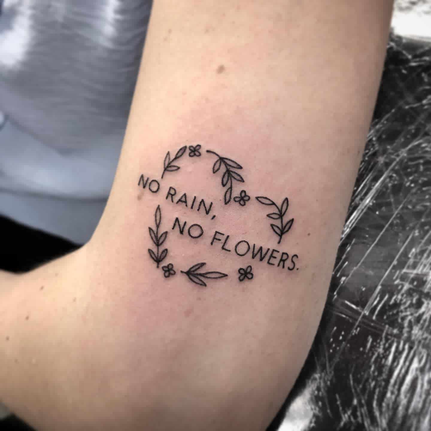 Tattoo with a floral design such as flowers and leaves encircling the text "No rain, no flowers"