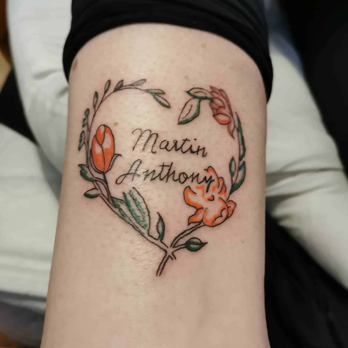 Tattoo of heart-shaped floral design with "Martin Anthony" written inside, featuring orange flowers and green leaves