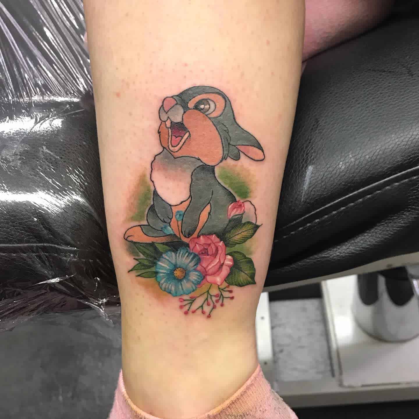 Tattoo of a cartoon rabbit sitting, surrounded by colorful flowers and green leaves