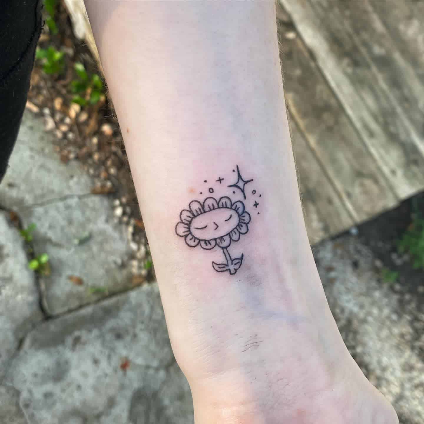 Cute black and grey tattoo of a smiling flower with a simple line drawing, accented with small sparkles and stars