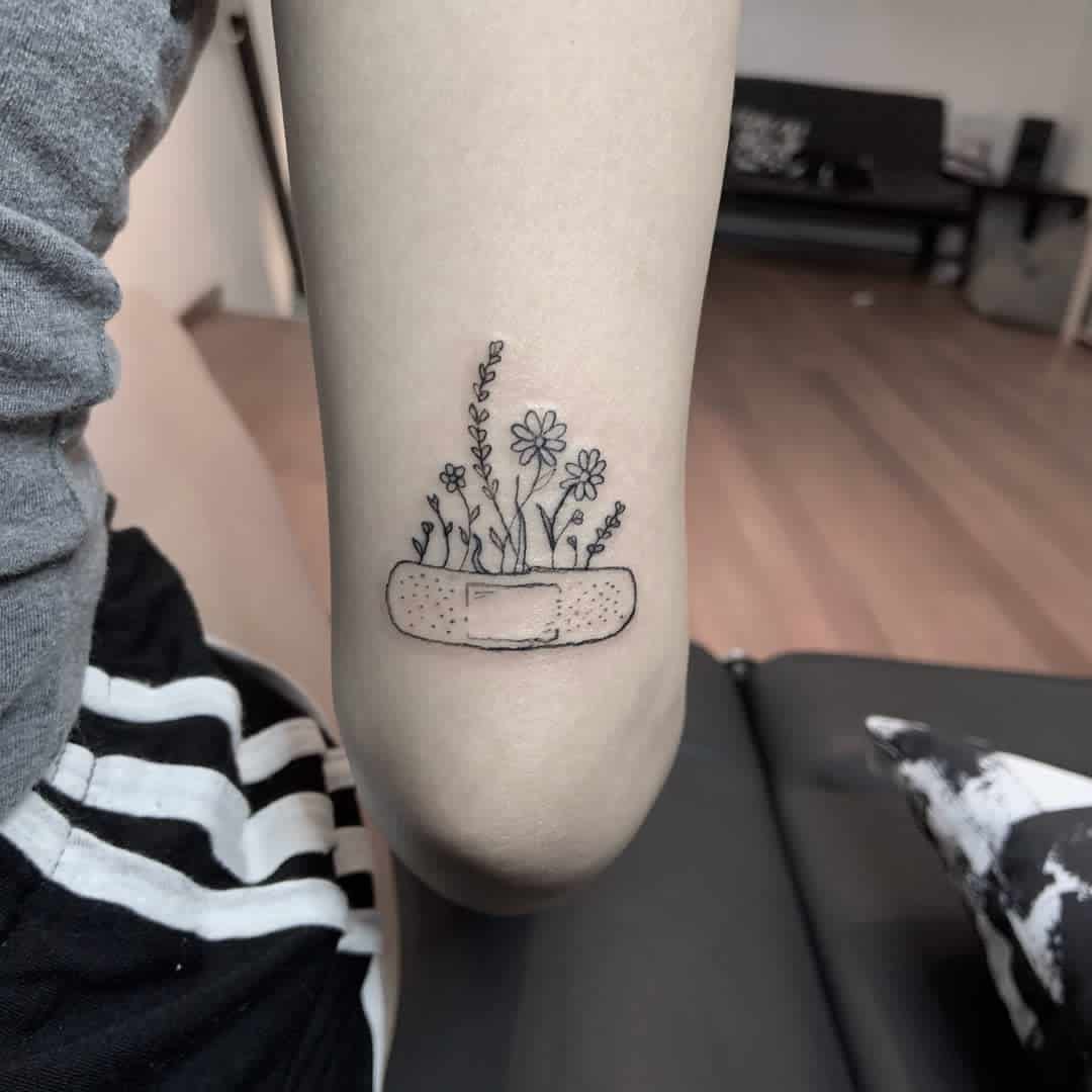 A tattoo shows a bandage with flowers growing from it, featuring daisies and other small blooms in black ink