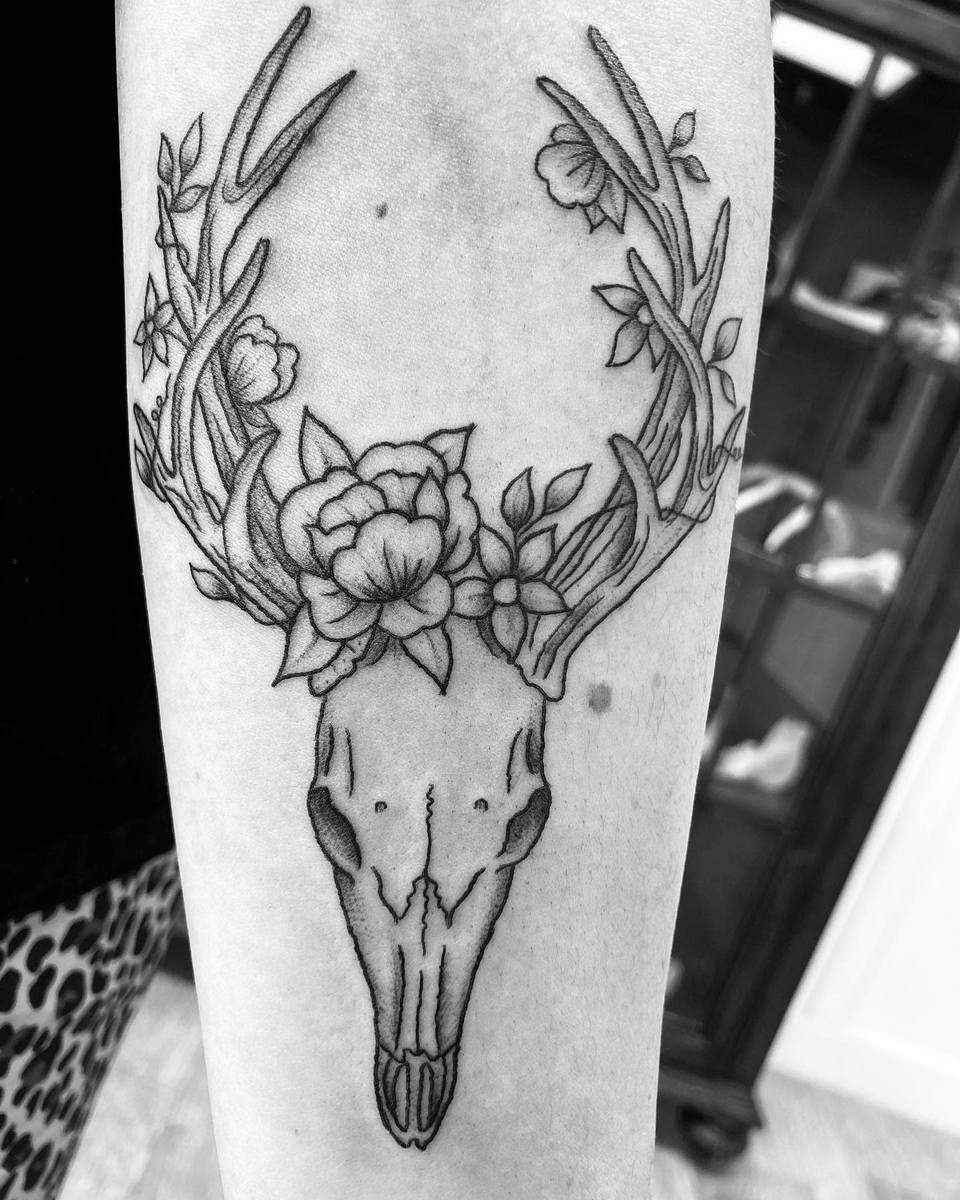 Black and white tattoo of a deer skull with antlers adorned by flowers on a forearm