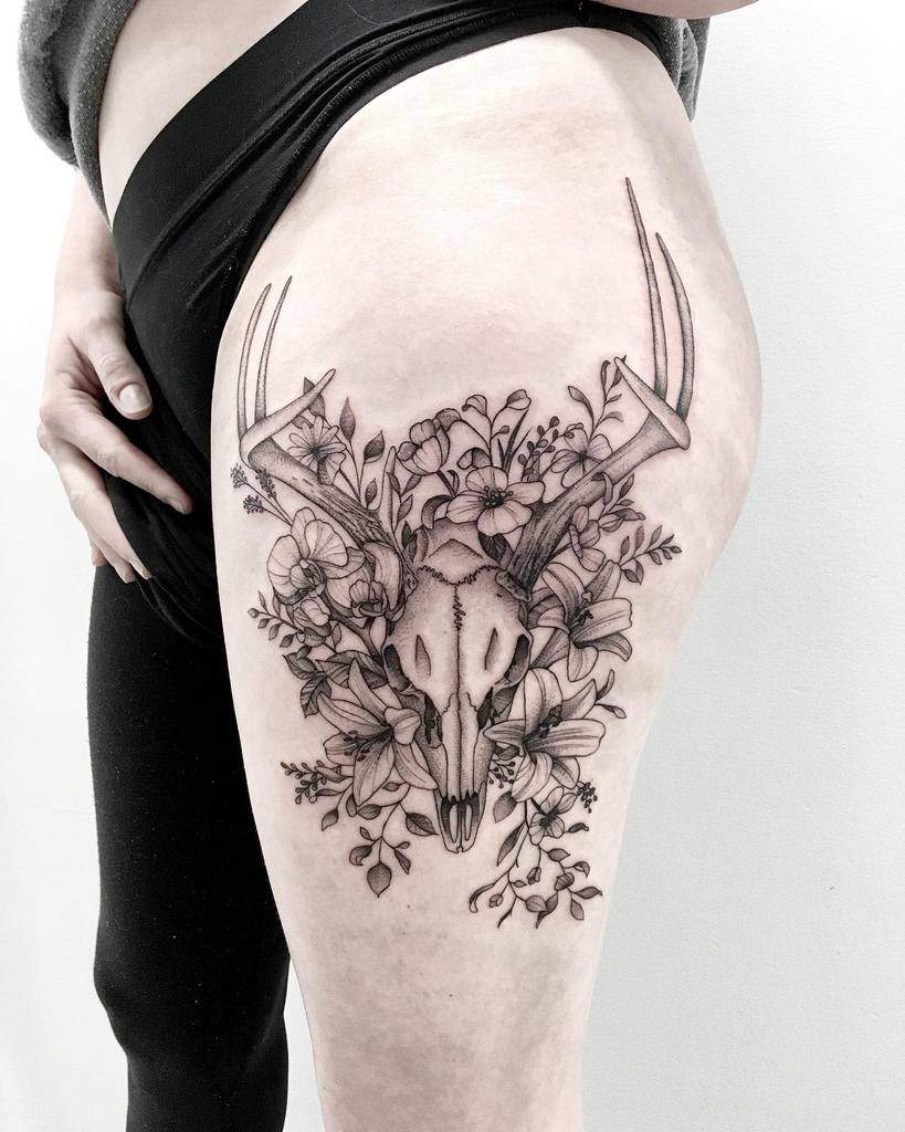 A thigh tattoo featuring a delicate deer skull nestled among vibrant flowers and intricate branches