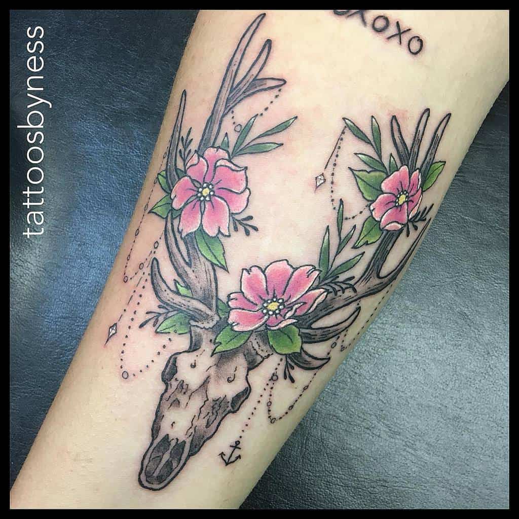 Tattoo of a deer skull with antlers adorned with pink flowers and green leaves on a person's arm, showcasing a harmonious blend of nature and rugged elegance