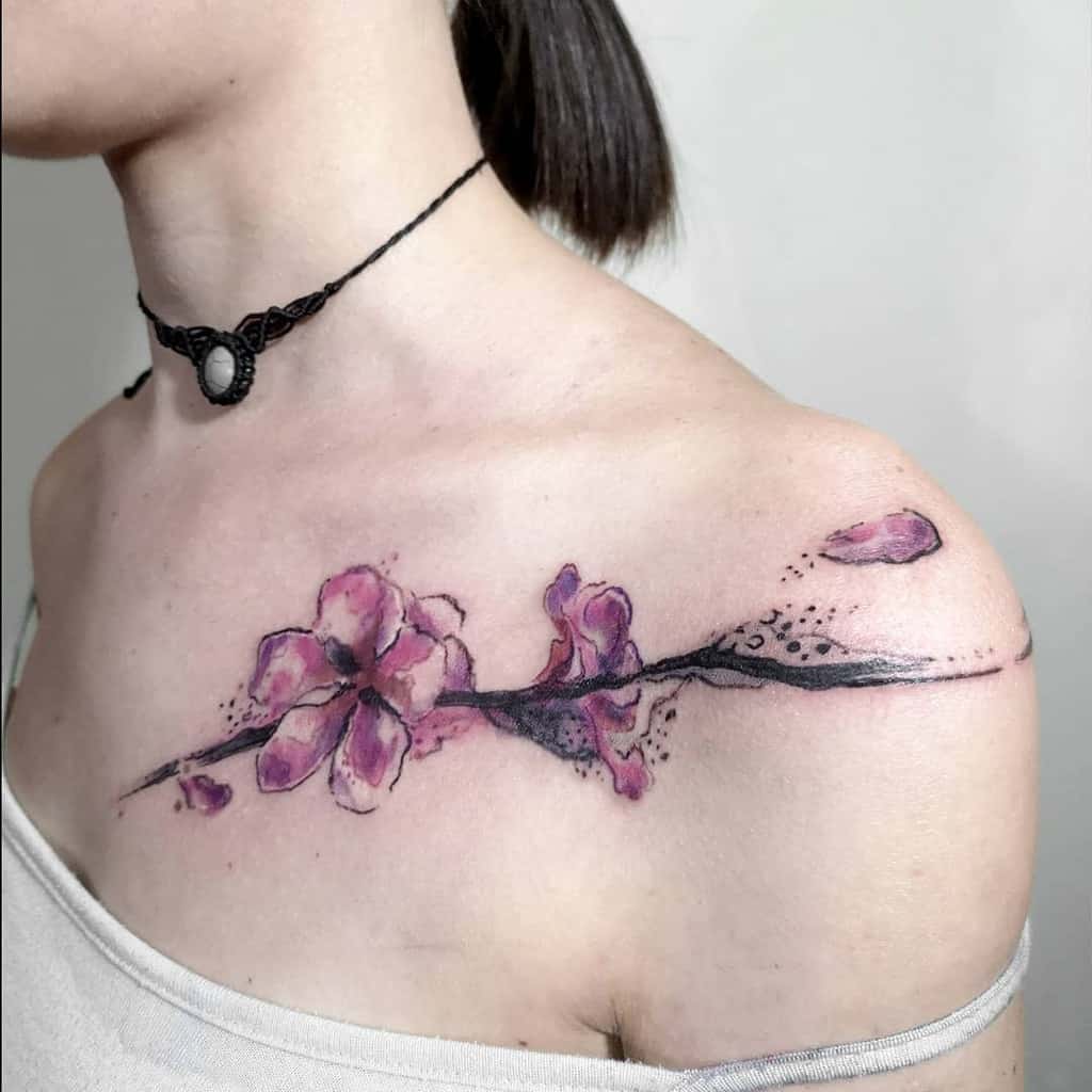Artistic watercolor-style tattoo of a cherry blossom branch stretching across the collarbone, with delicate pink petals and ink splatter details