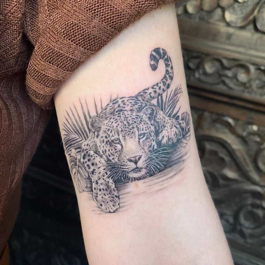 A realistic black and grey tattoo of a jaguar crouching in the grass, with a focus on its calm expression and detailed fur.
