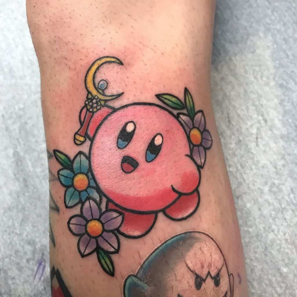 Kirby tattoo holding a crescent moon staff, surrounded by colorful flowers and leaves