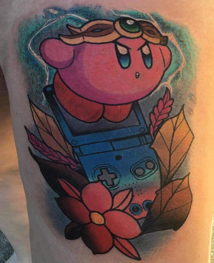 Kirby tattoo standing on a Game Boy Advance, wearing a mystical headpiece, surrounded by flowers and leaves with a glowing aura effect
