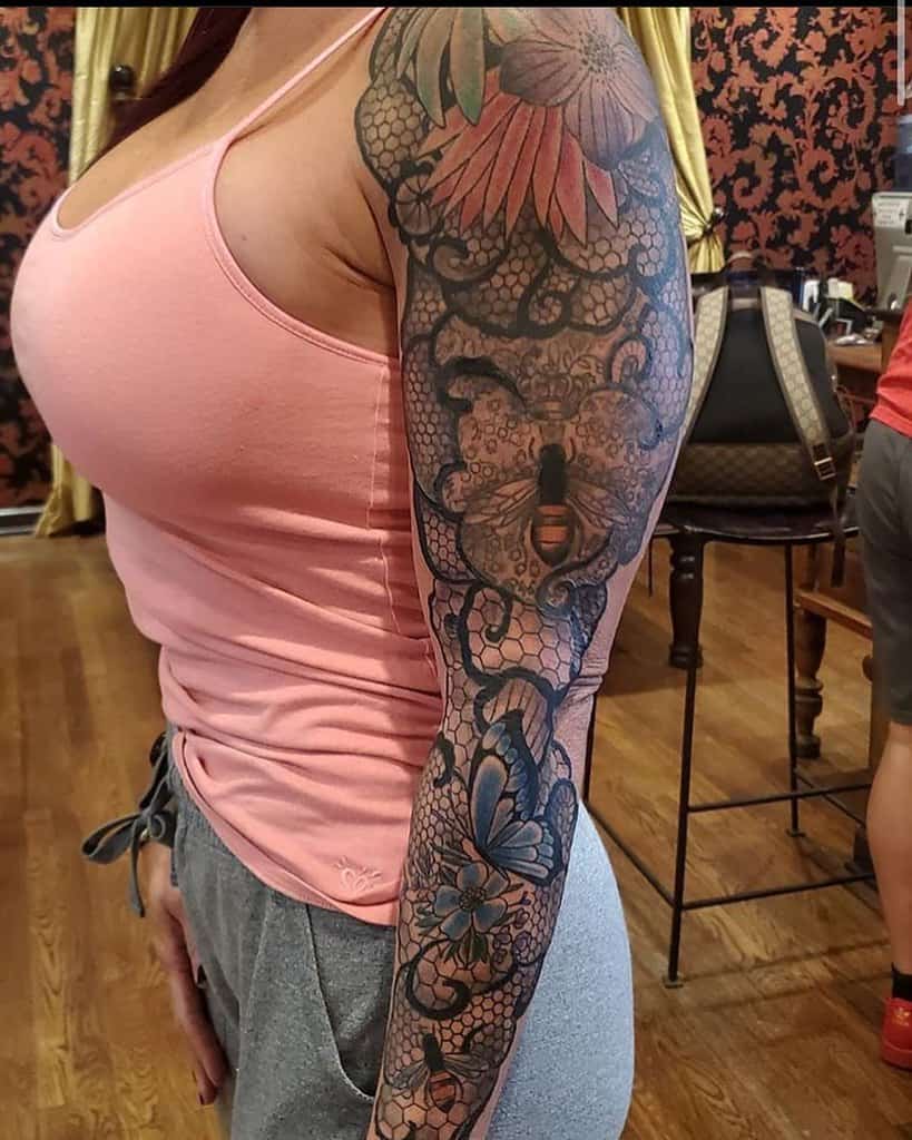 Elegant lace tattoo sleeve with floral details, a bee centerpiece, and butterfly accents, blending black and colored ink for a bold look.